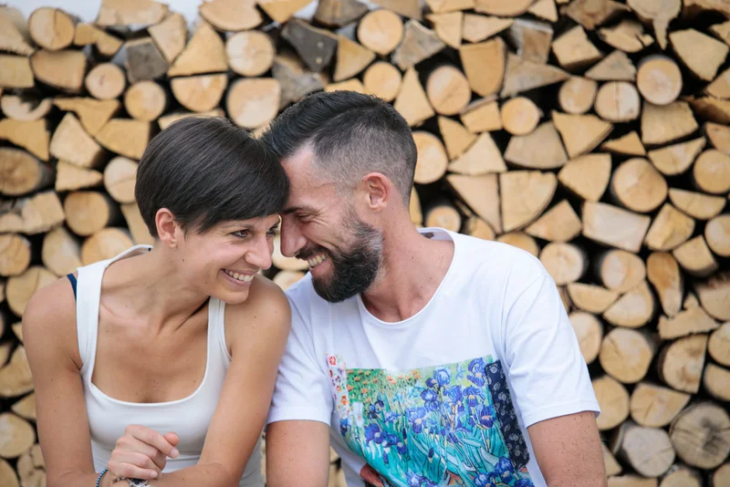 Coppia sorridente si scambia sguardi affettuosi davanti a un muro di legna.