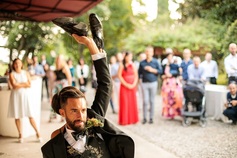 Sposo solleva una scarpa durante il ricevimento, mentre gli invitati osservano divertiti.