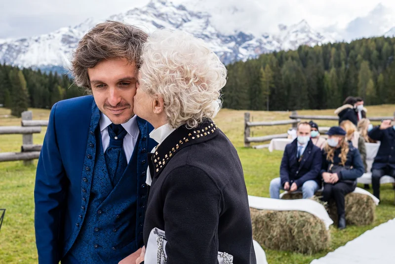 Una coppia si scambia un gesto affettuoso durante la cerimonia all'aperto in un rifugio a Cortina, con montagne sullo sfondo.
