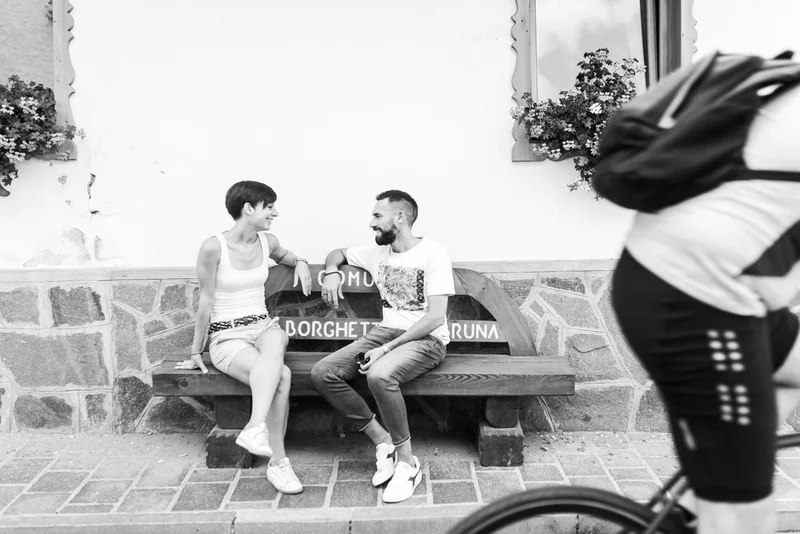 Coppia seduta su una panchina mentre chiacchiera, con un ciclista in movimento sullo sfondo.