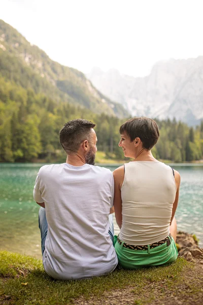 Coppia seduta sulla riva del lago, immersa nella natura, si scambia sguardi affettuosi.