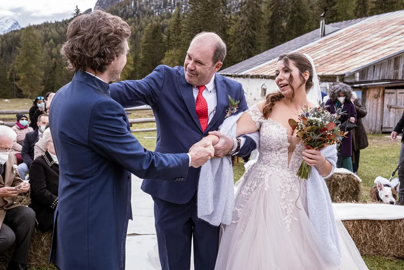 Sposi si scambiano un sorriso e una stretta di mano durante la cerimonia all'aperto a Cortina, circondati da invitati.
