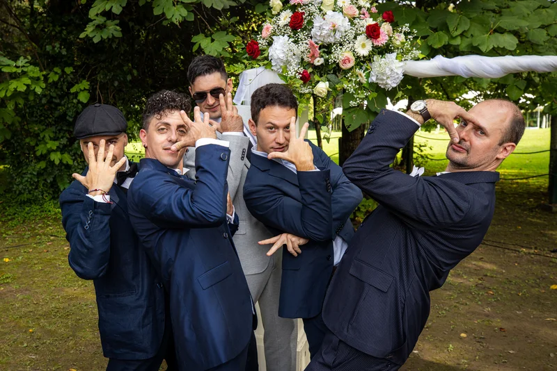 Gruppo di invitati in abiti eleganti che posano divertendosi sotto un arco floreale in un parco.