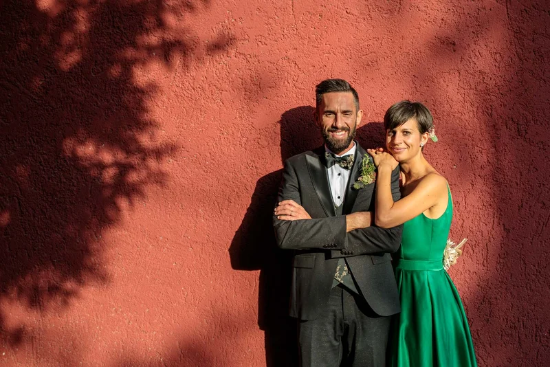 Coppia sorridente in posa davanti a un muro rosso, con ombre che creano un'atmosfera calda.