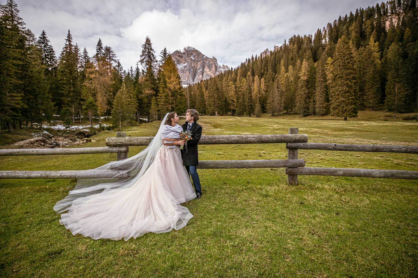 Coppia di sposi si abbraccia in un prato verde con alberi e montagne sullo sfondo.